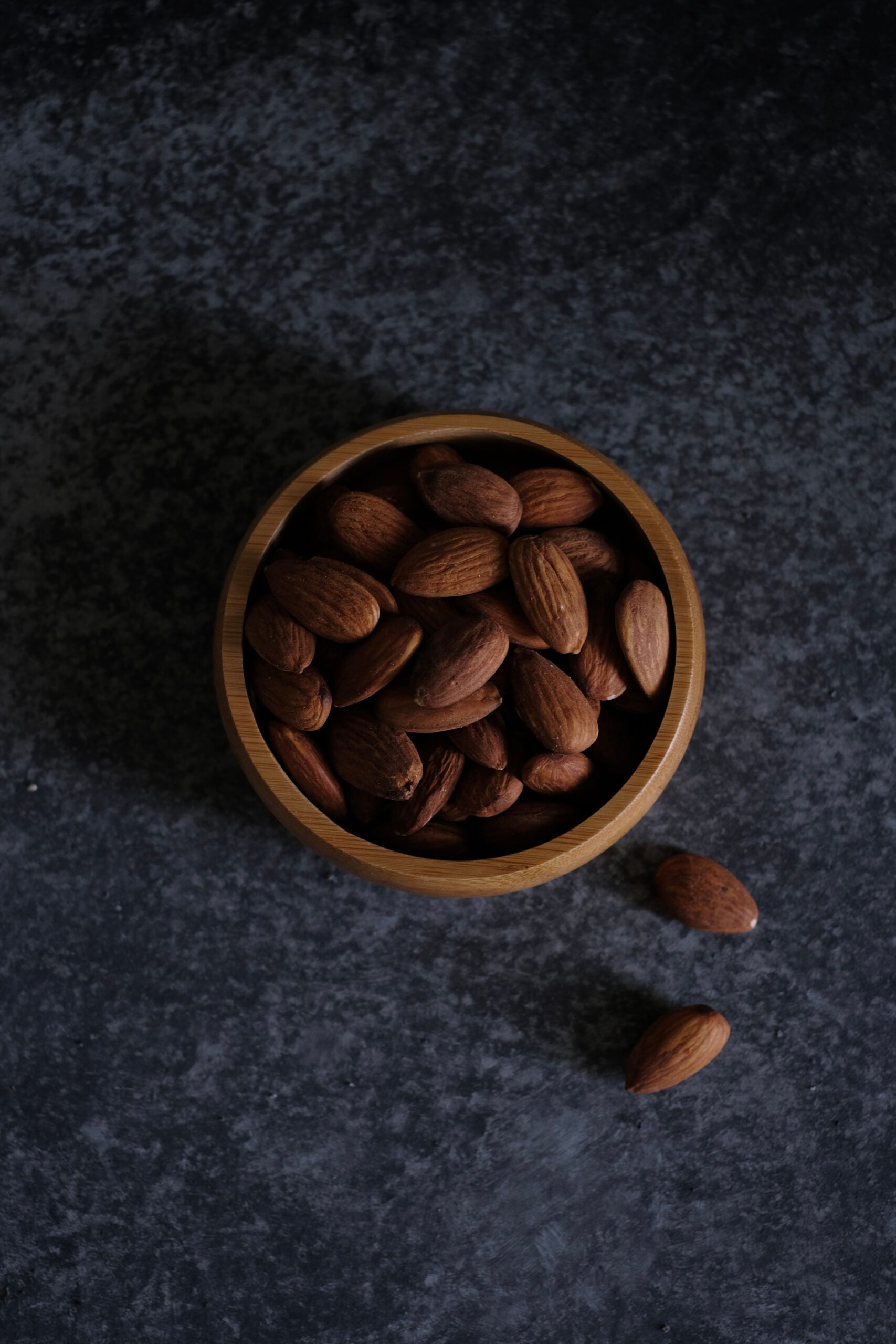 Close-up of almonds in a wooden bowl, highlighting healthy snacking options on a dark textured surface.